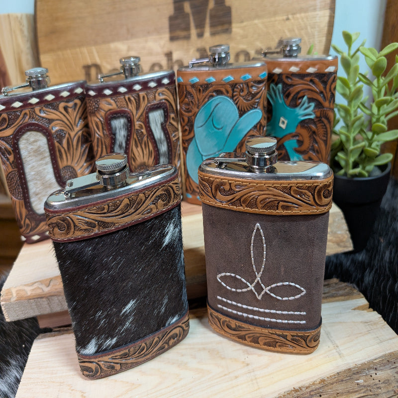 Two leather cowhide flasks with tooled designs, one with a cap and one without, displayed on a wooden surface.