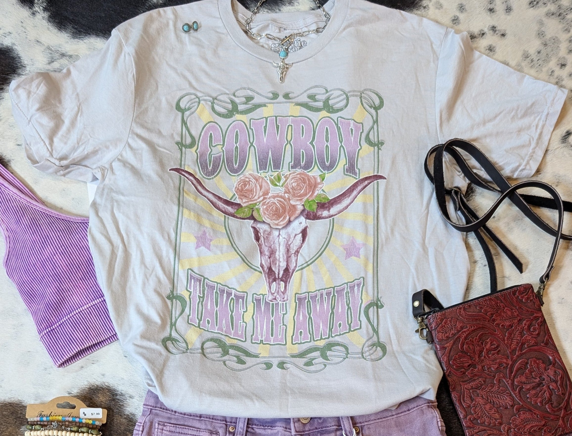 “Flat lay of Cowboy Take Me Away graphic tee by Goodie Two Sleeves with purple shorts, western fringe purse, and accessories.”