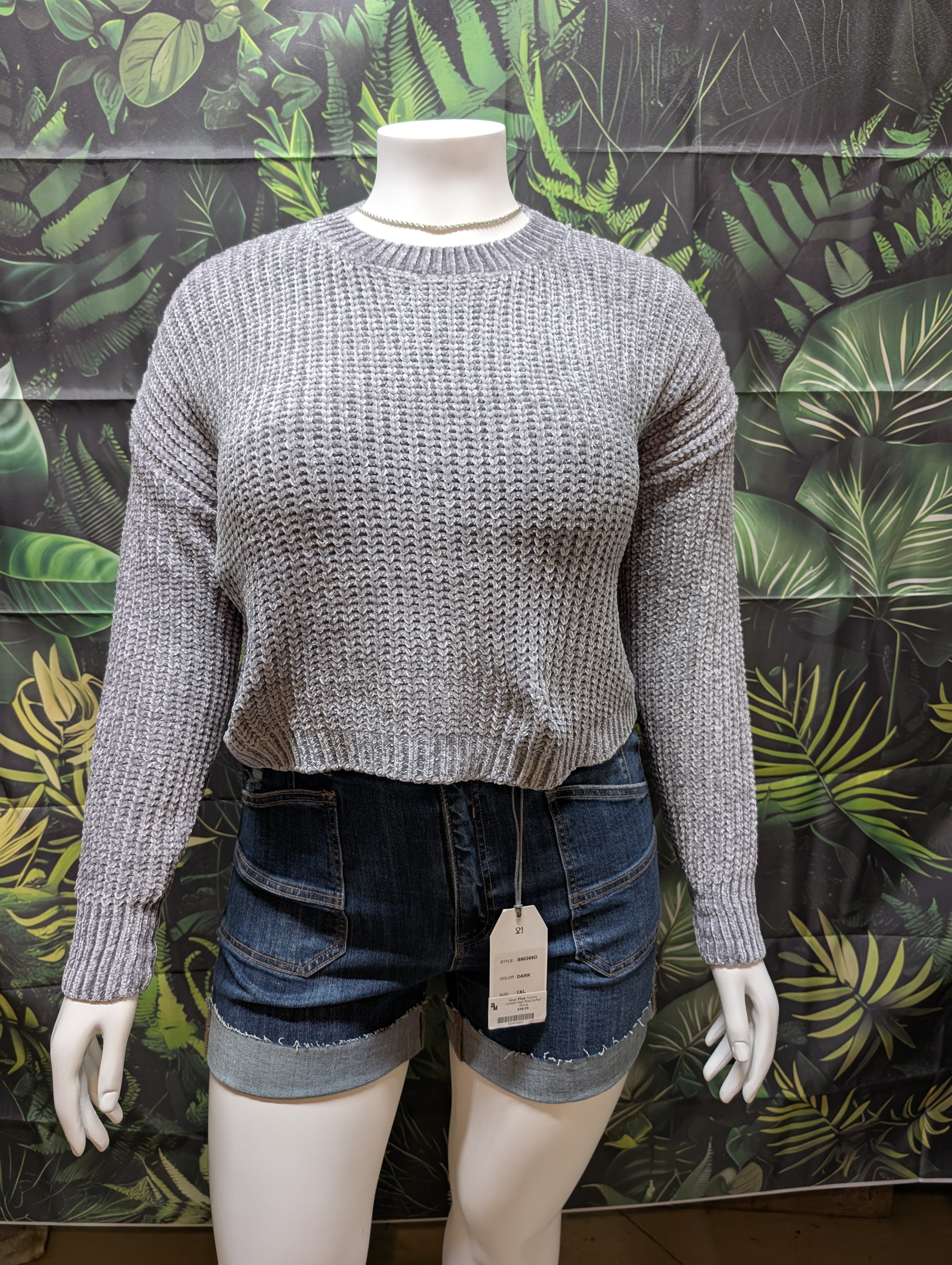 Mannequin wearing a gray sweater and denim shorts with a tag against a leaf-patterned wall.