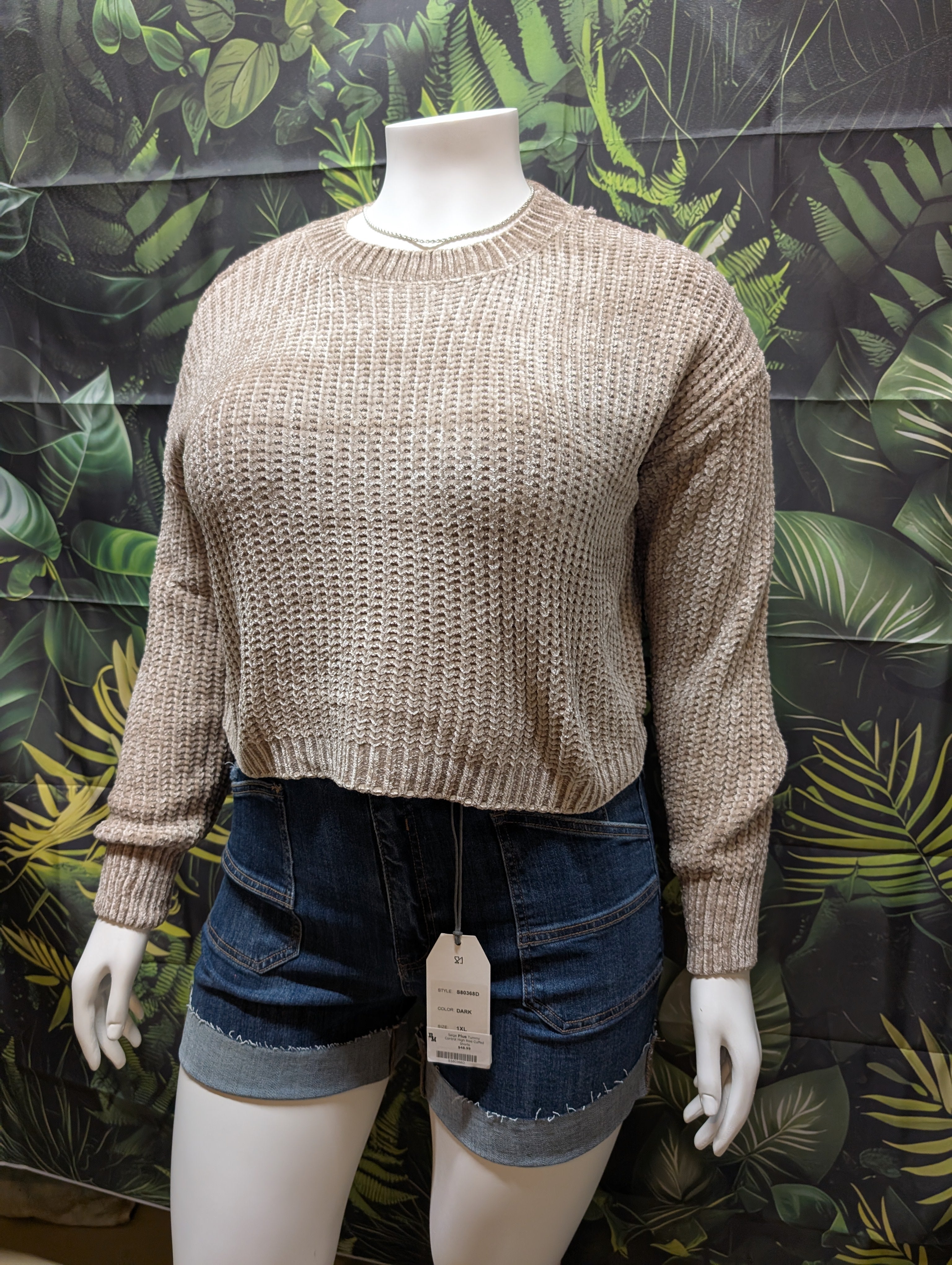 Mannequin wearing a beige sweater and denim shorts with a green leafy background