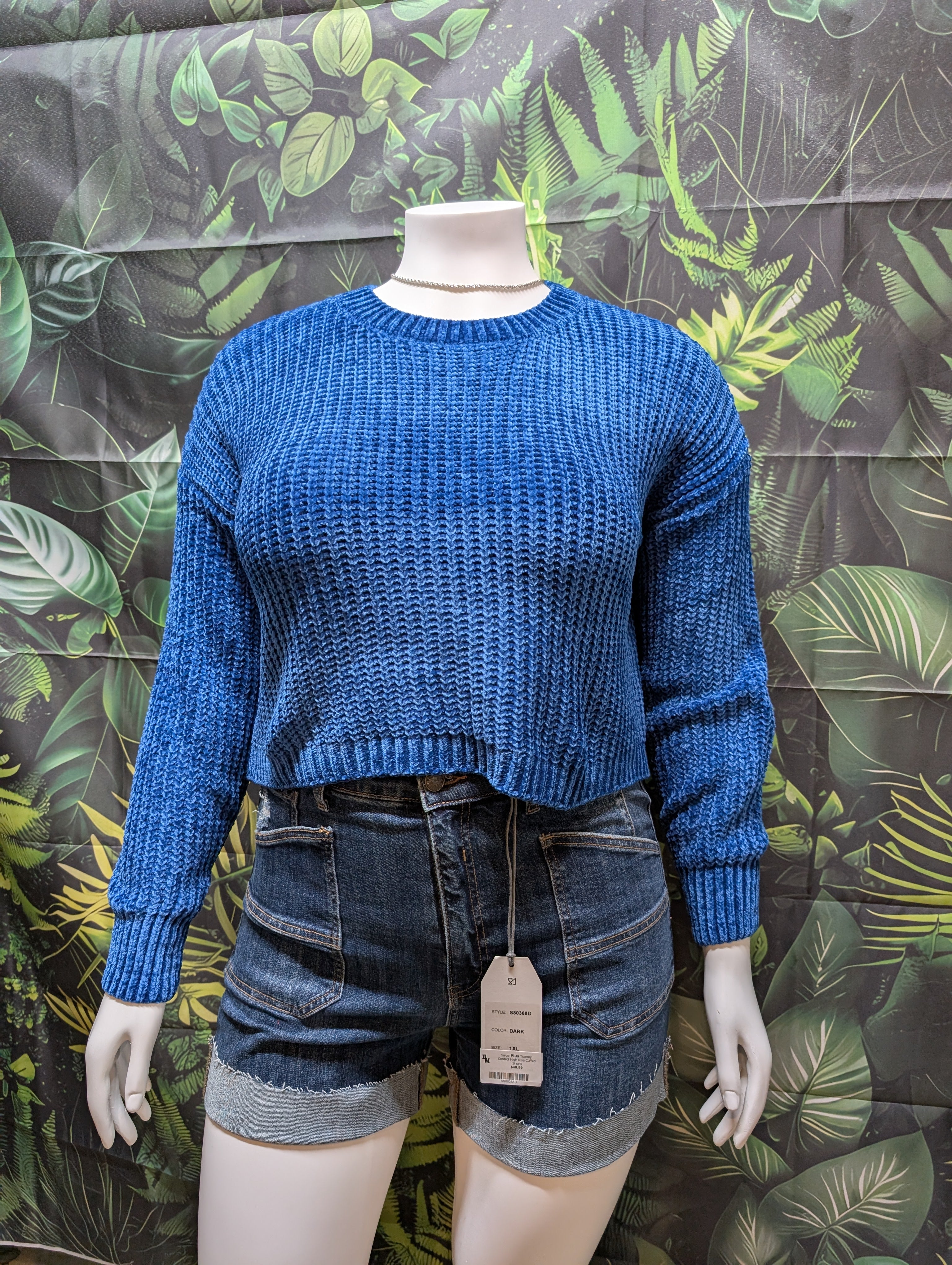 Mannequin wearing a blue sweater and denim shorts with a leaf-patterned background