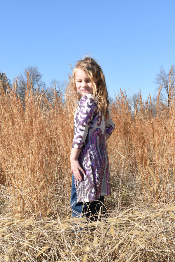 Young girl standing in brown grass wearing a western purple and white duster.