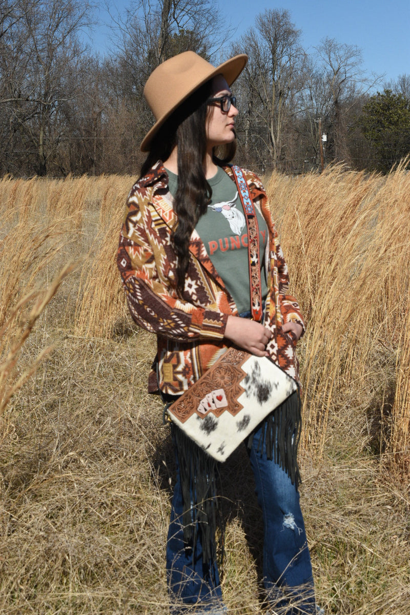 Lady looking at the sun with a hat and a western aztec shacket and a cowhide purse with playing cards on it.