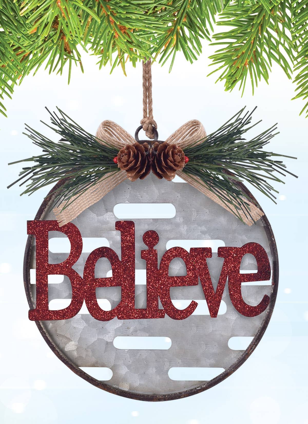 A metal Christmas ornament with the word "Believe" in glittery red letters, hanging from pine branches with a burlap bow and two pinecones.