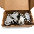 Four industrial caster wheels with gray rubber tires and silver metal swivel housings, shown inside an open brown cardboard box .