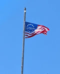 America's 250th Anniversary 3x5 flag in classic red, white, and blue flying on a flagpole against a blue sky.