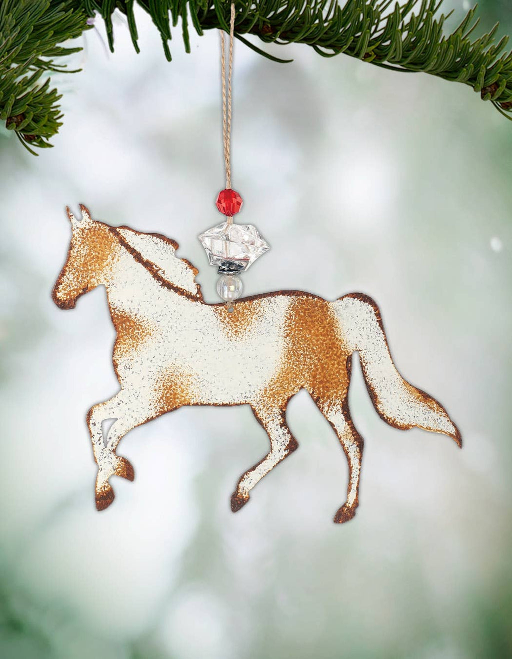 A horse-shaped Christmas ornament hanging from a pine branch. The ornament has a textured white and brown speckled design with a small red bead and clear faceted crystal charm.