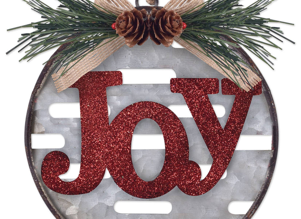 A round rustic Christmas ornament featuring the word 'Joy' in glittery red letters over a white slatted background, decorated with pine branches, a burlap ribbon, and two pinecones at the top.