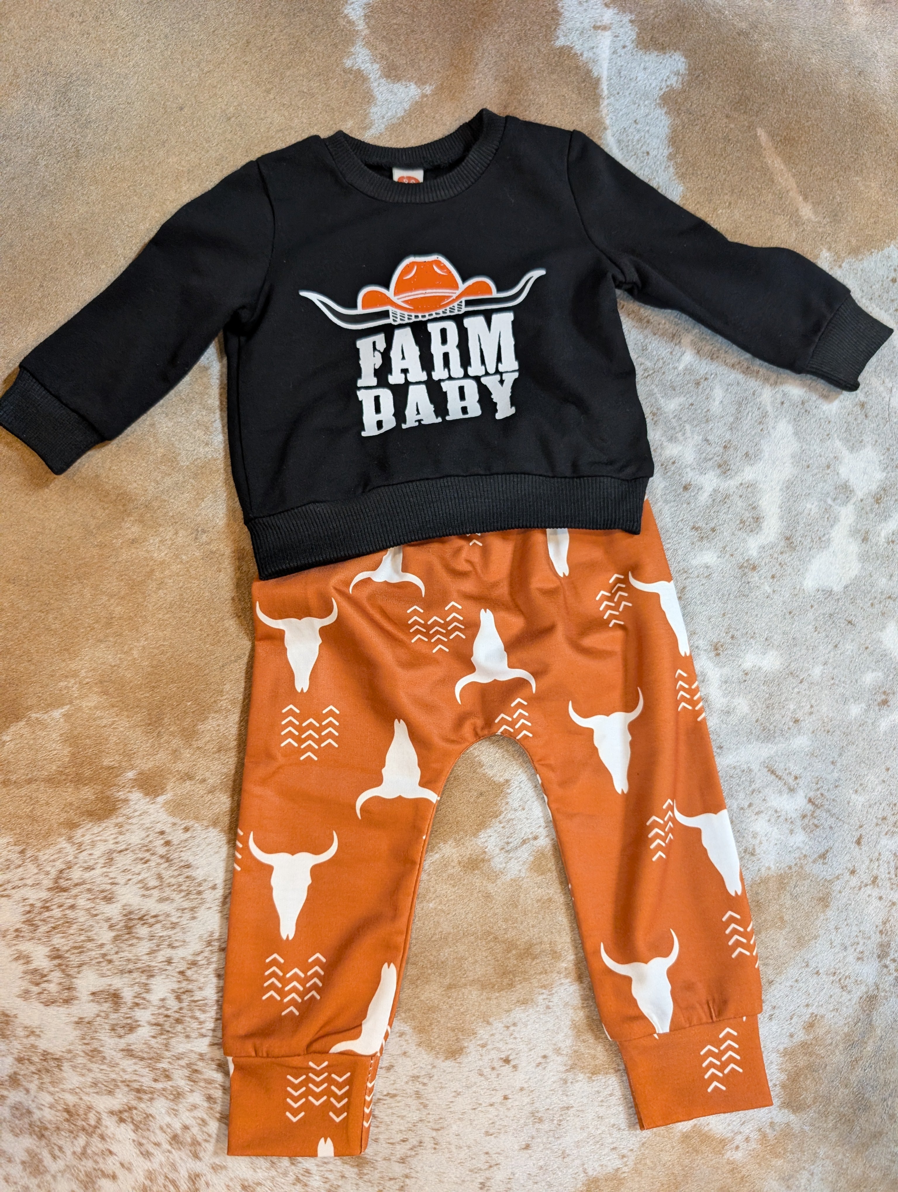 Black sweatshirt with 'Farm Baby' text and orange pants with longhorn bull pattern on a textured surface.