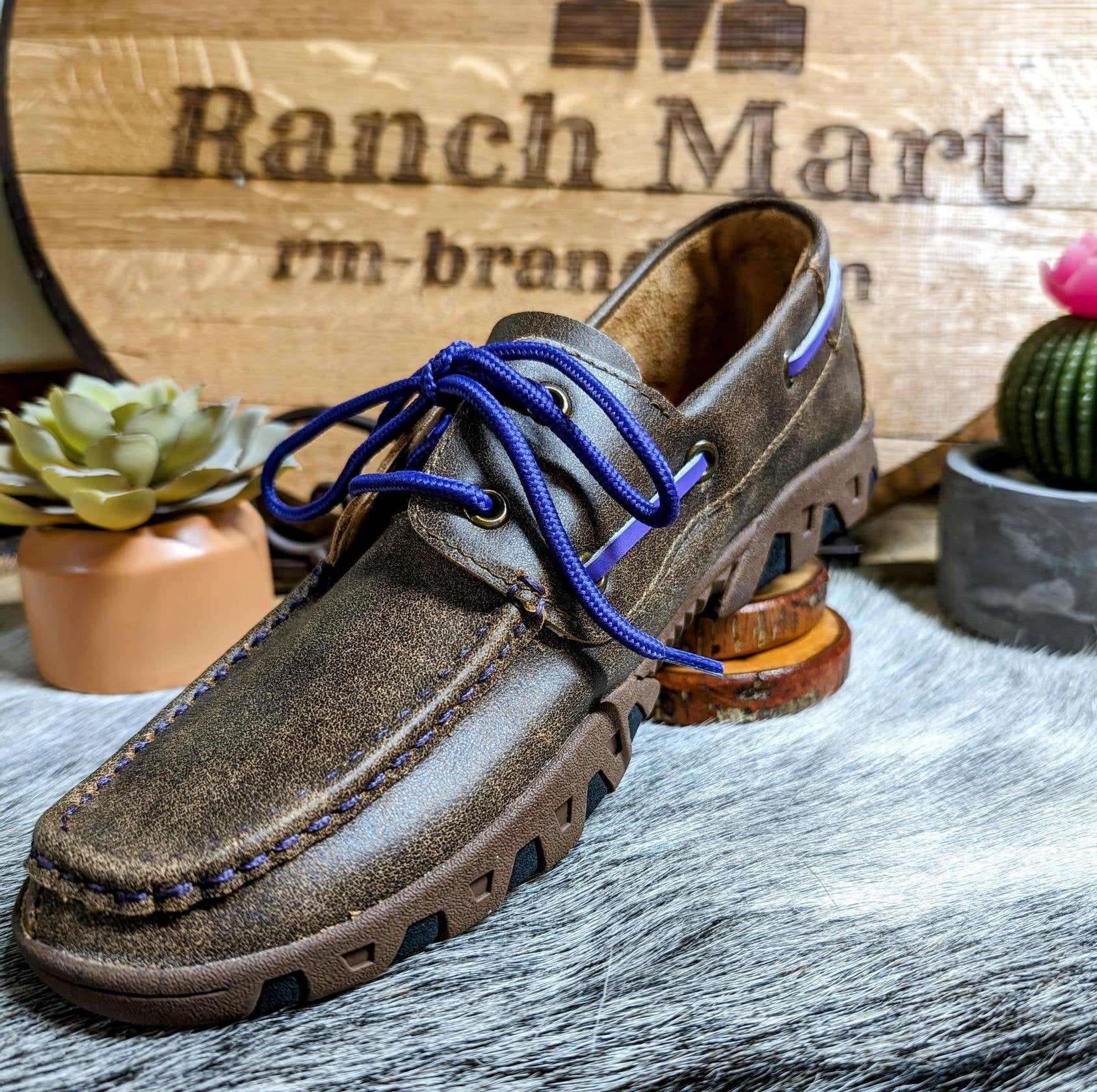 A Ferrini women's loafer with distressed brown leather and purple laces, displayed on a wooden surface with plants in the background.