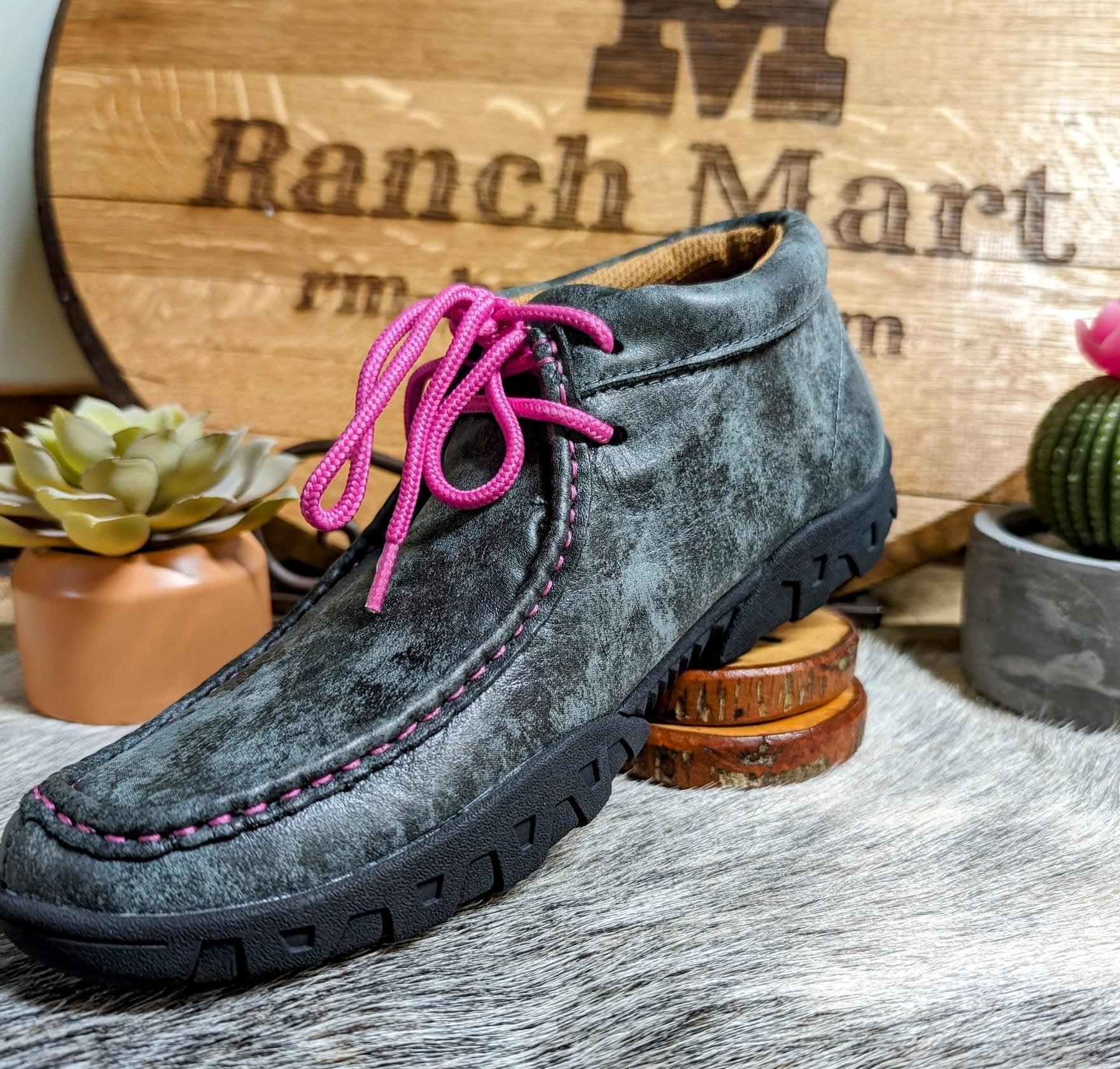 A Ferrini women's Rogue ankle shoe with distressed black leather and bright pink laces, displayed on a wooden surface with plants and a cactus in the background.