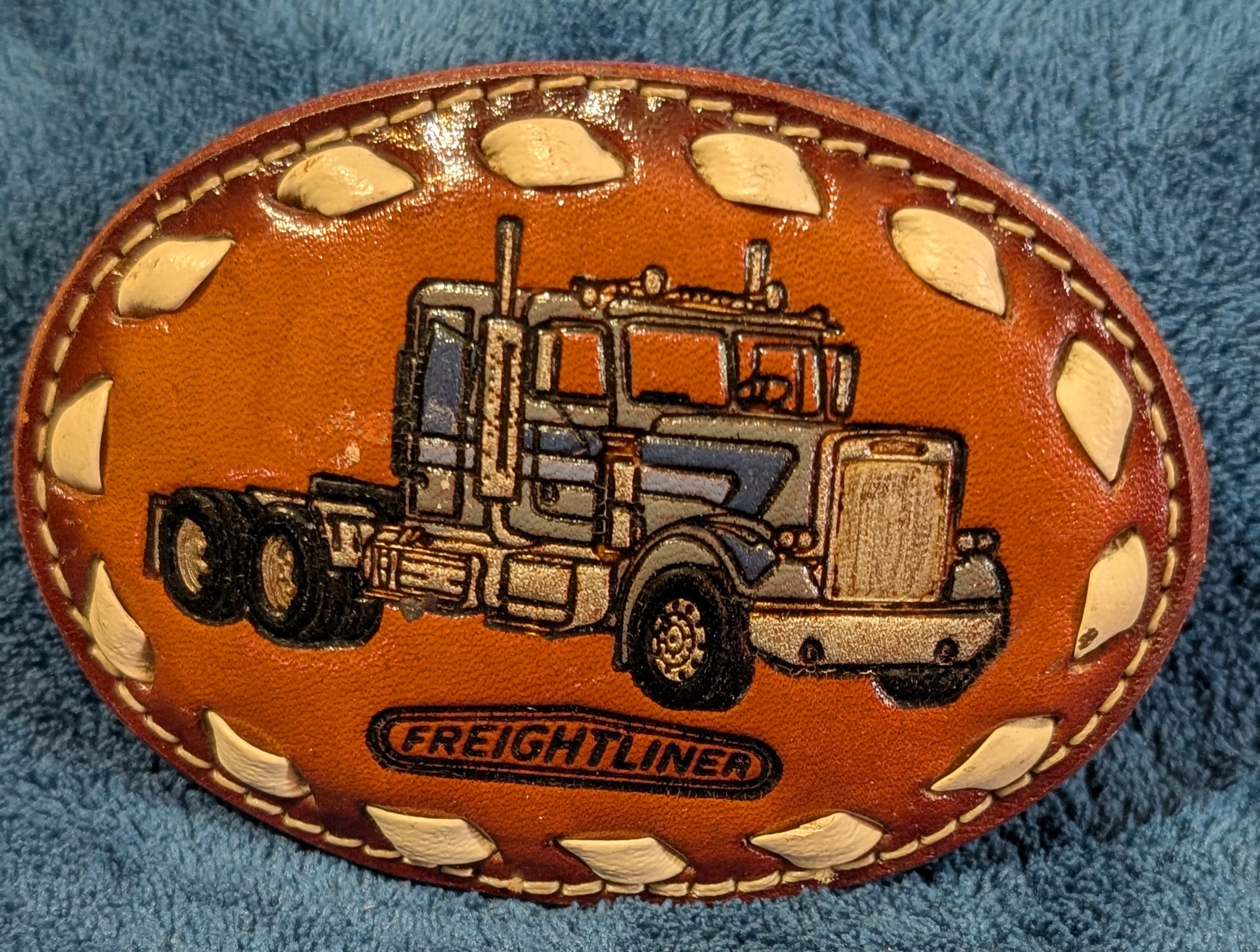 Leather belt buckle with truck design and 'Freightliner' branding on a blue fabric background