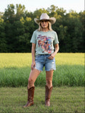 Light green t-shirt with graphic design on a lady with cowboy hat & boots in a outdoor setting.