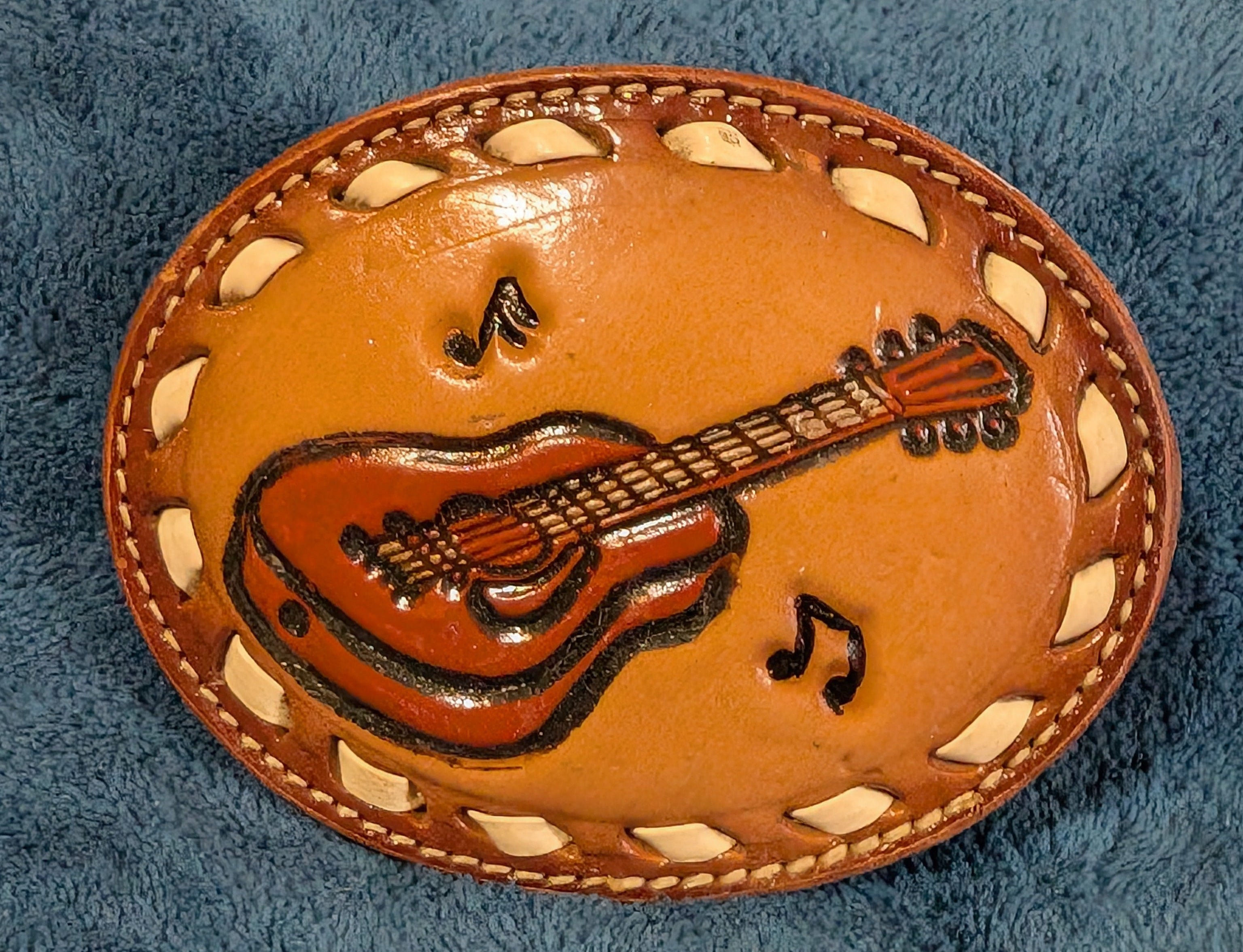 Brown leather belt buckle with guitar and music note design on a blue fabric background