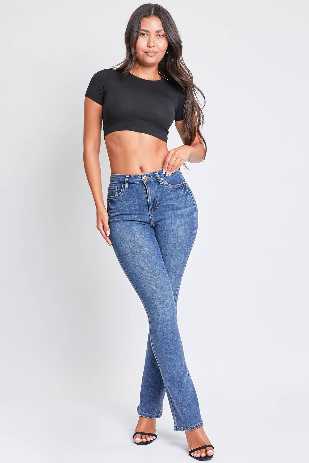A woman standing, wearing ultra high-rise bootcut jeans in a medium wash blue denim color, with a black cropped top and black strappy sandals.