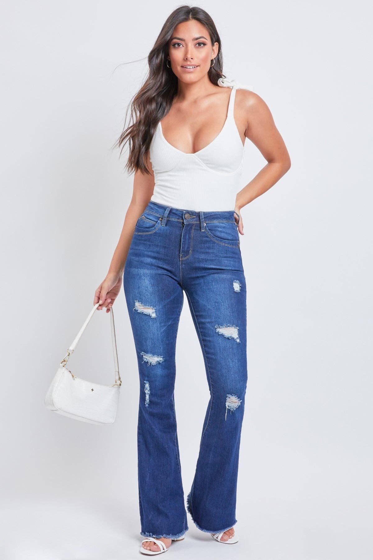 A woman wearing a white tank top and high-rise flare jeans with a frayed hem and light whiskering. She has a white handbag and is posing against a neutral background.