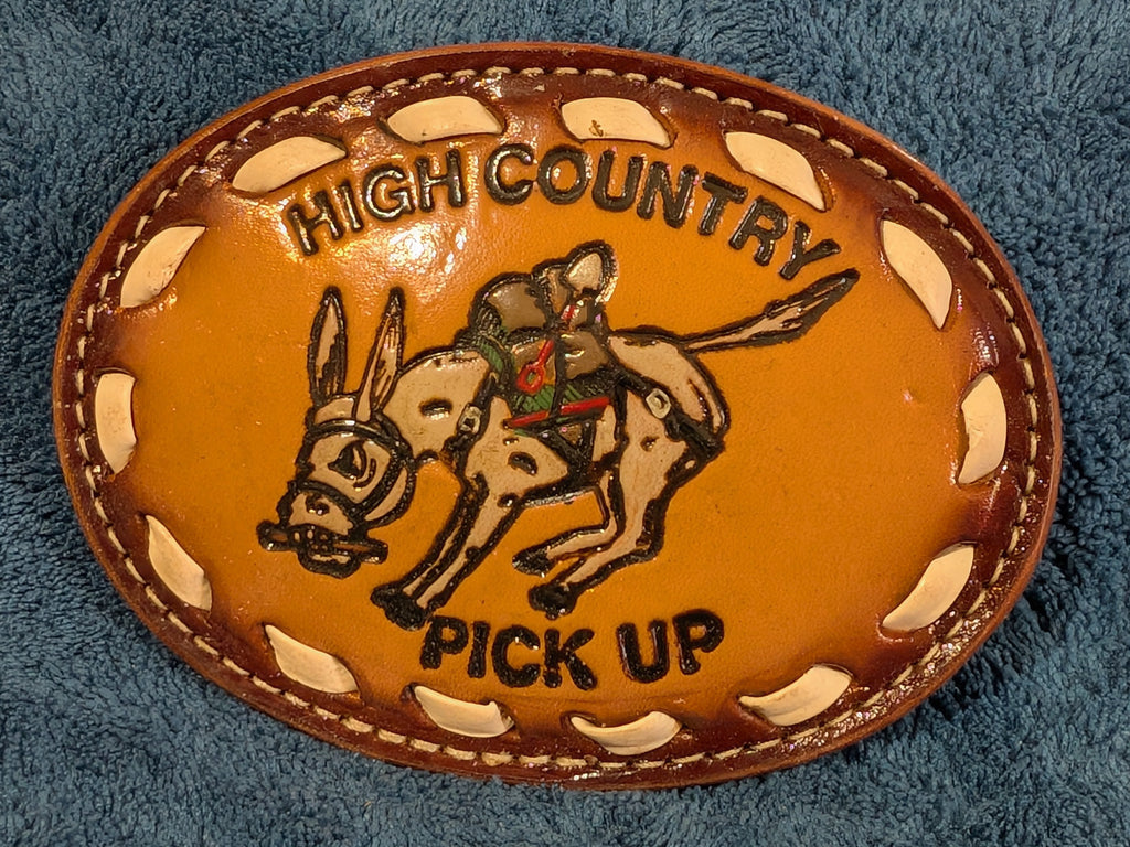 Leather belt buckle with donkey design and 'High Country Pick Up' text on a blue textured background
