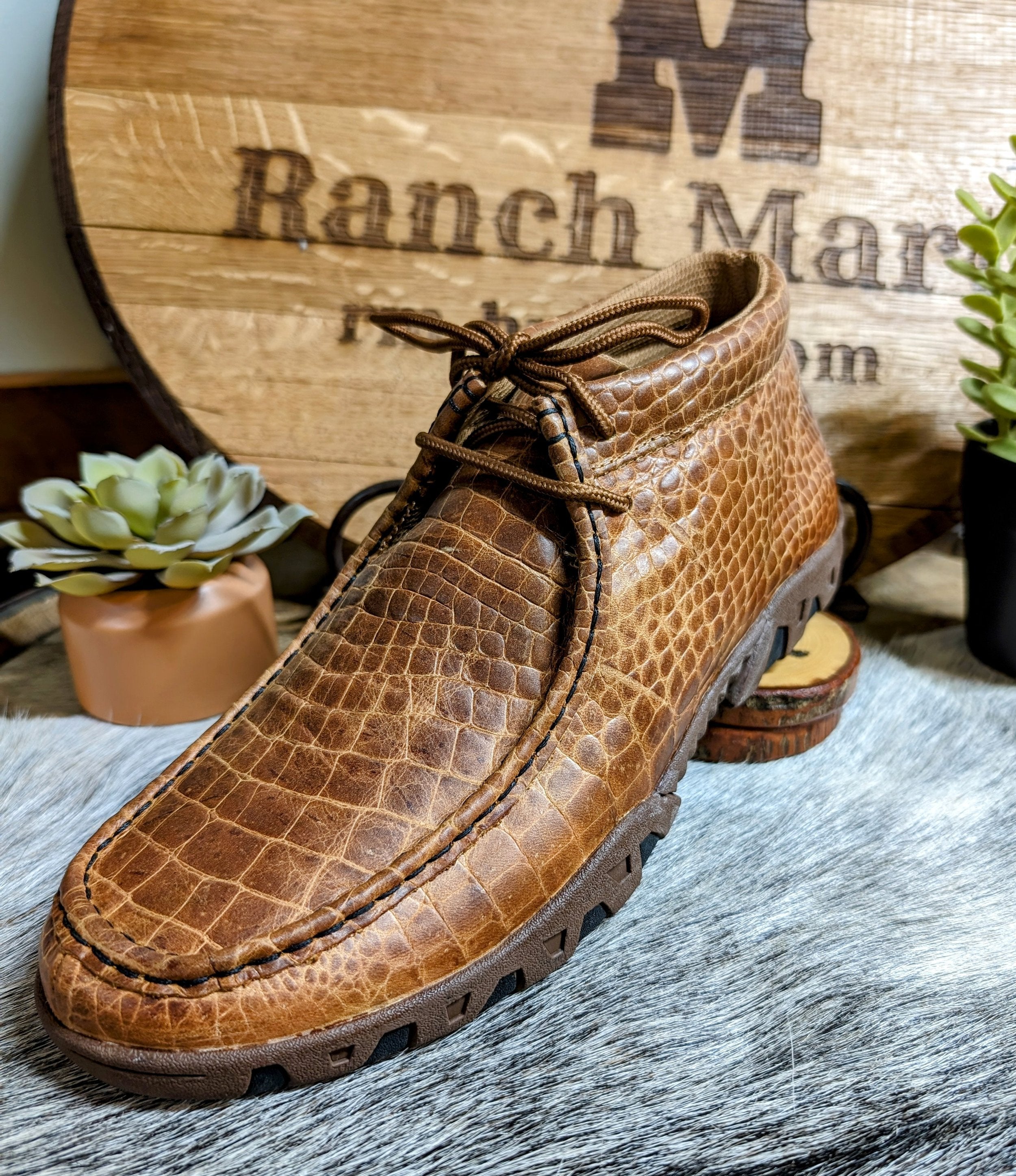 A brown Ferrini men's shoe with an alligator belly cowhide print displayed on a wooden surface, alongside a wooden sign with 'Ranch Mart' branding in the background.