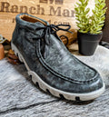 A Ferrini Men's Rogue Ankle Shoe in smokey black color, displayed against a rustic background with a plant and wooden objects.