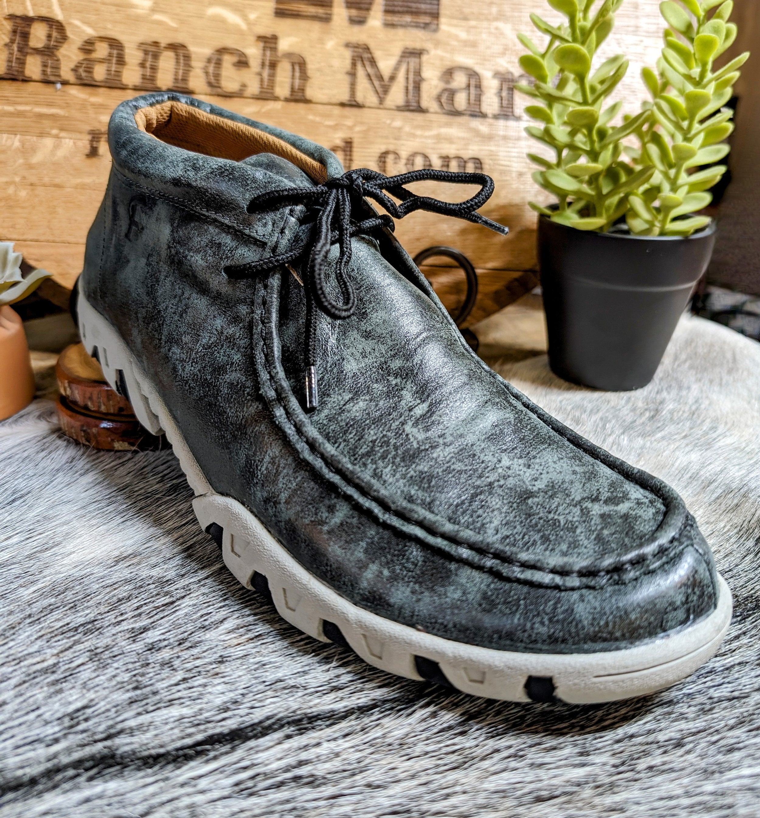 A Ferrini Men's Rogue Ankle Shoe in smokey black color, displayed against a rustic background with a plant and wooden objects.