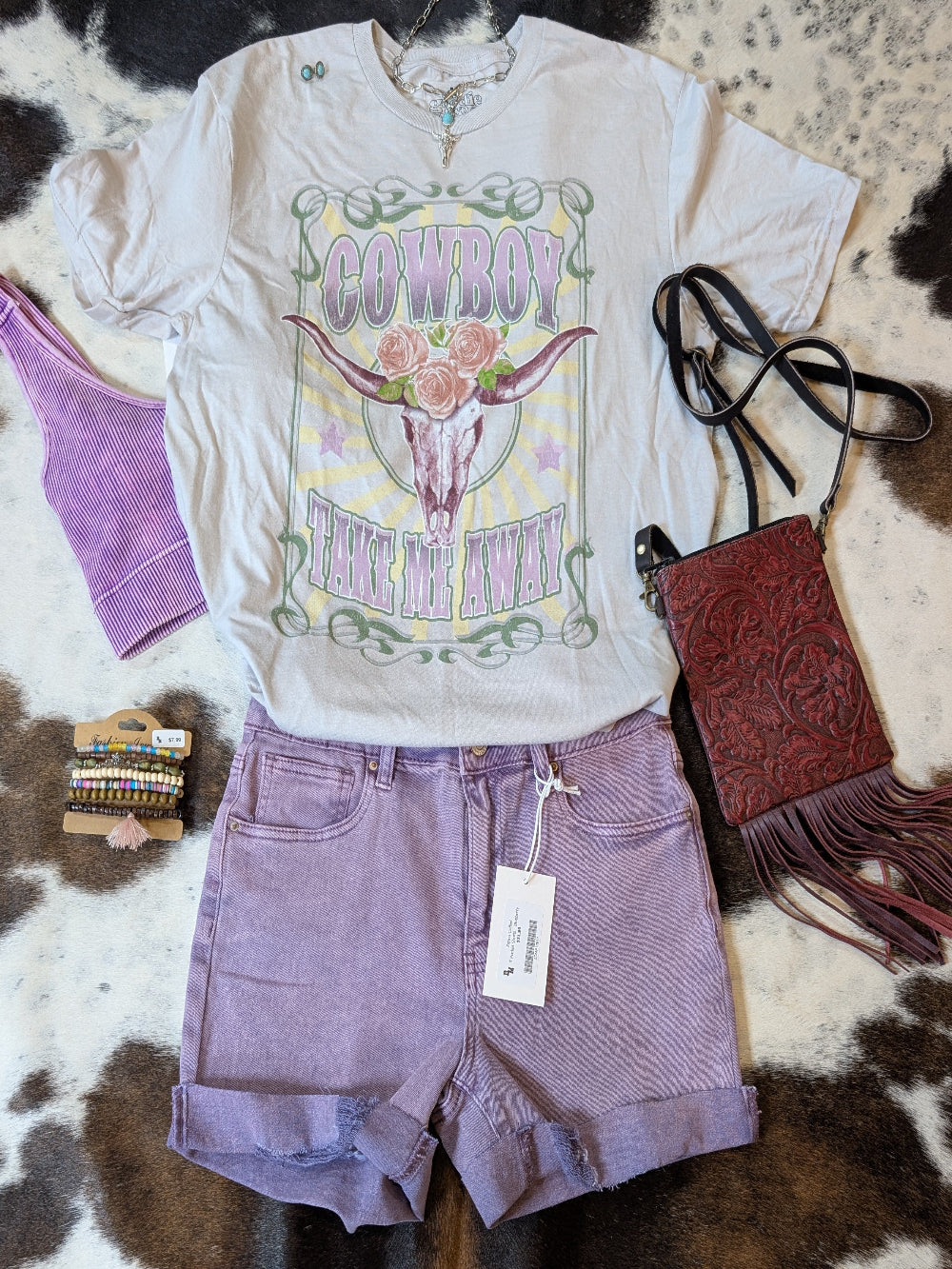 “Goodie Two Sleeves Cowboy Take Me Away graphic tee styled with lilac denim shorts, fringe western purse, and boho jewelry on cowhide background.”