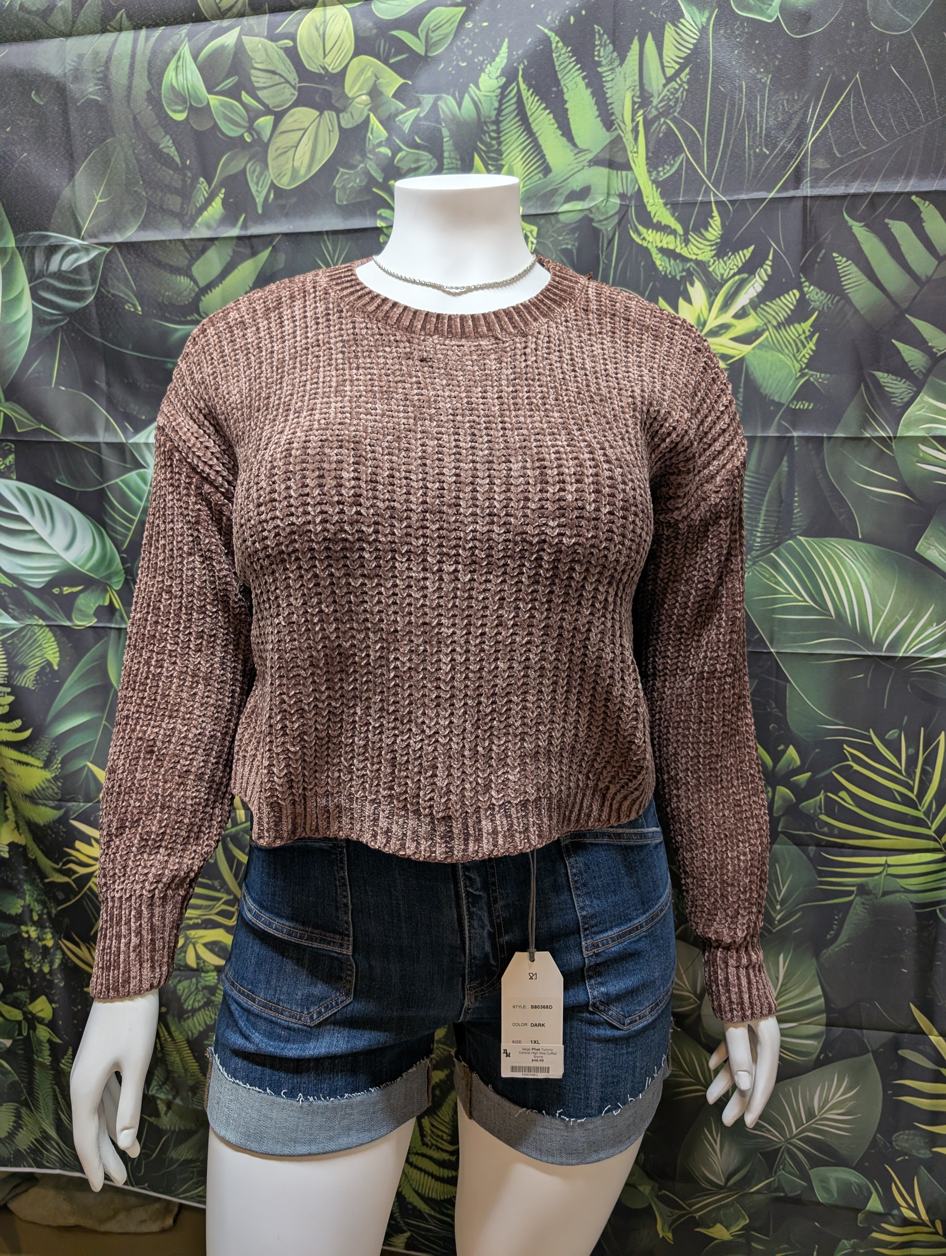 Mannequin wearing a brown knitted sweater and denim shorts against a leaf-patterned background