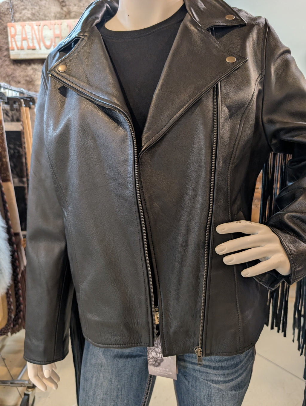 Mannequin wearing a black leather jacket with fringe details in a store setting.