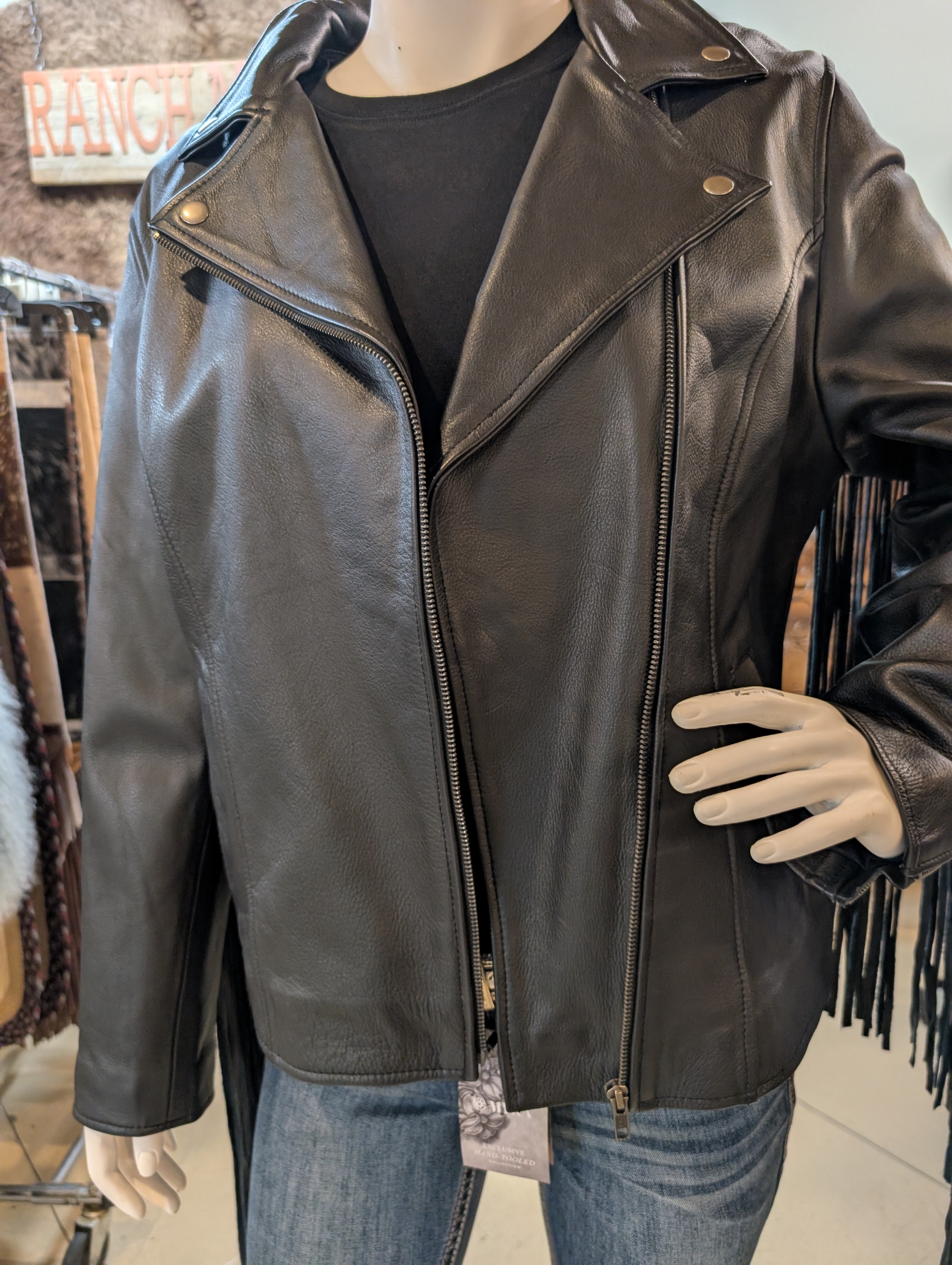 Mannequin wearing a black leather jacket with fringe details in a store setting.