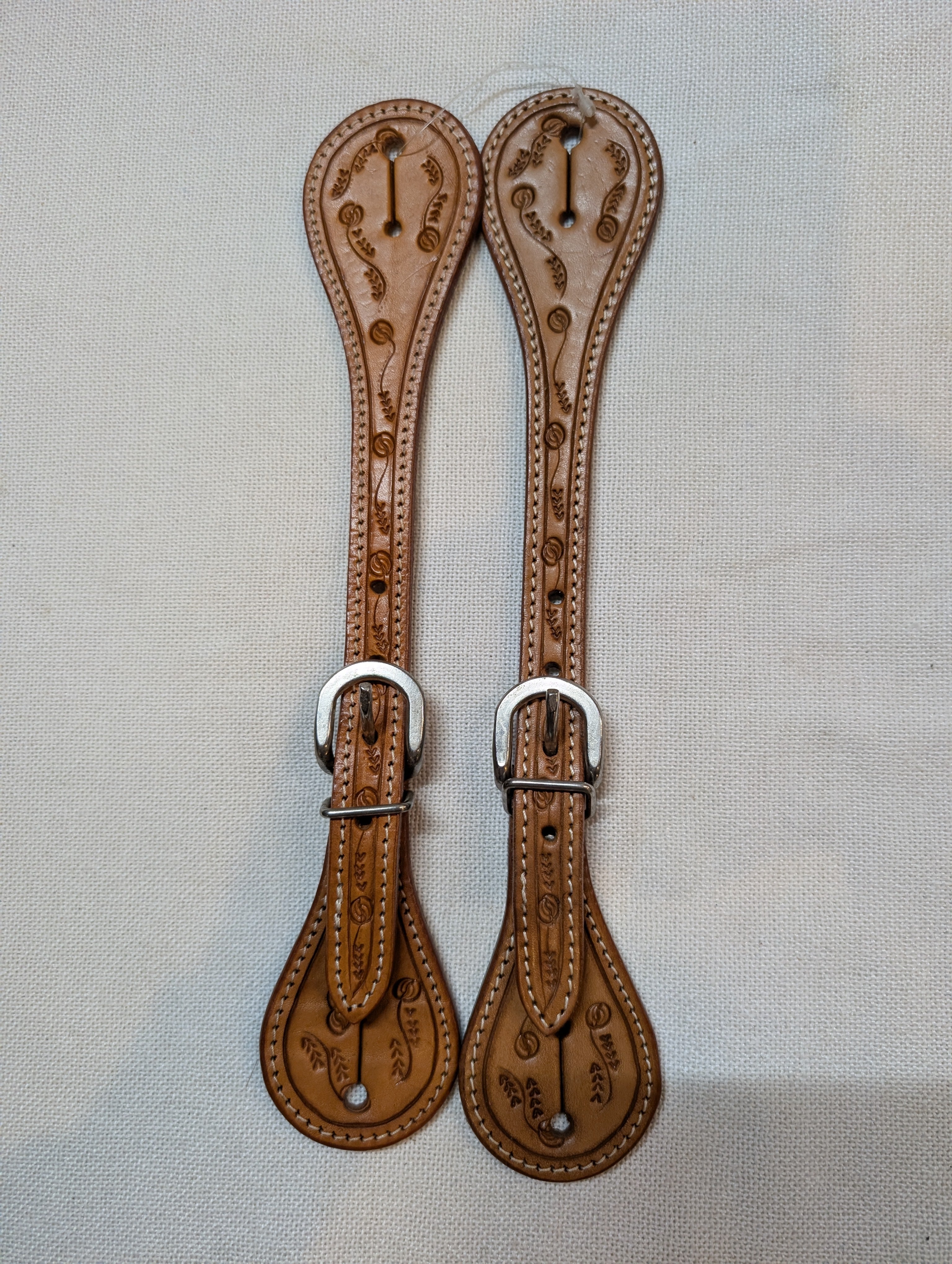 Pair of brown leather spurs with silver buckles on a light gray background