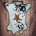 A star-shaped cowhide doormat with a brown star emblem on a white and black spotted background, displayed against a wooden backdrop.
