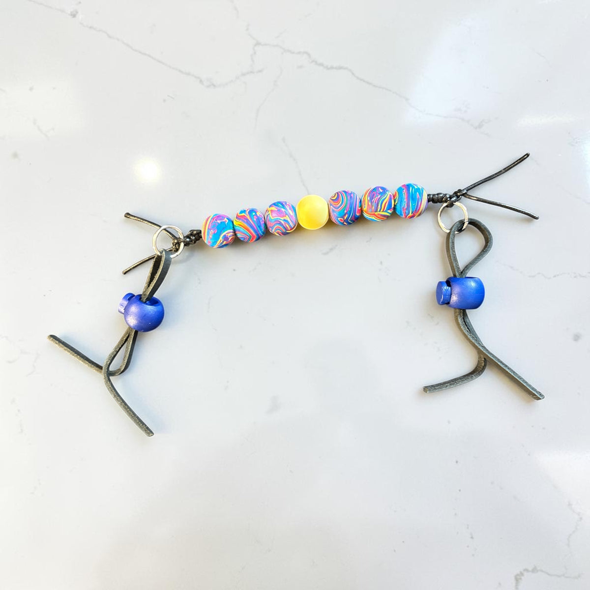 A handmade metal and bead ornament featuring two silver-toned stick-figure charms with blue beads, connected by a row of marbled beads in blue, pink, purple, and yellow on a white marble surface.