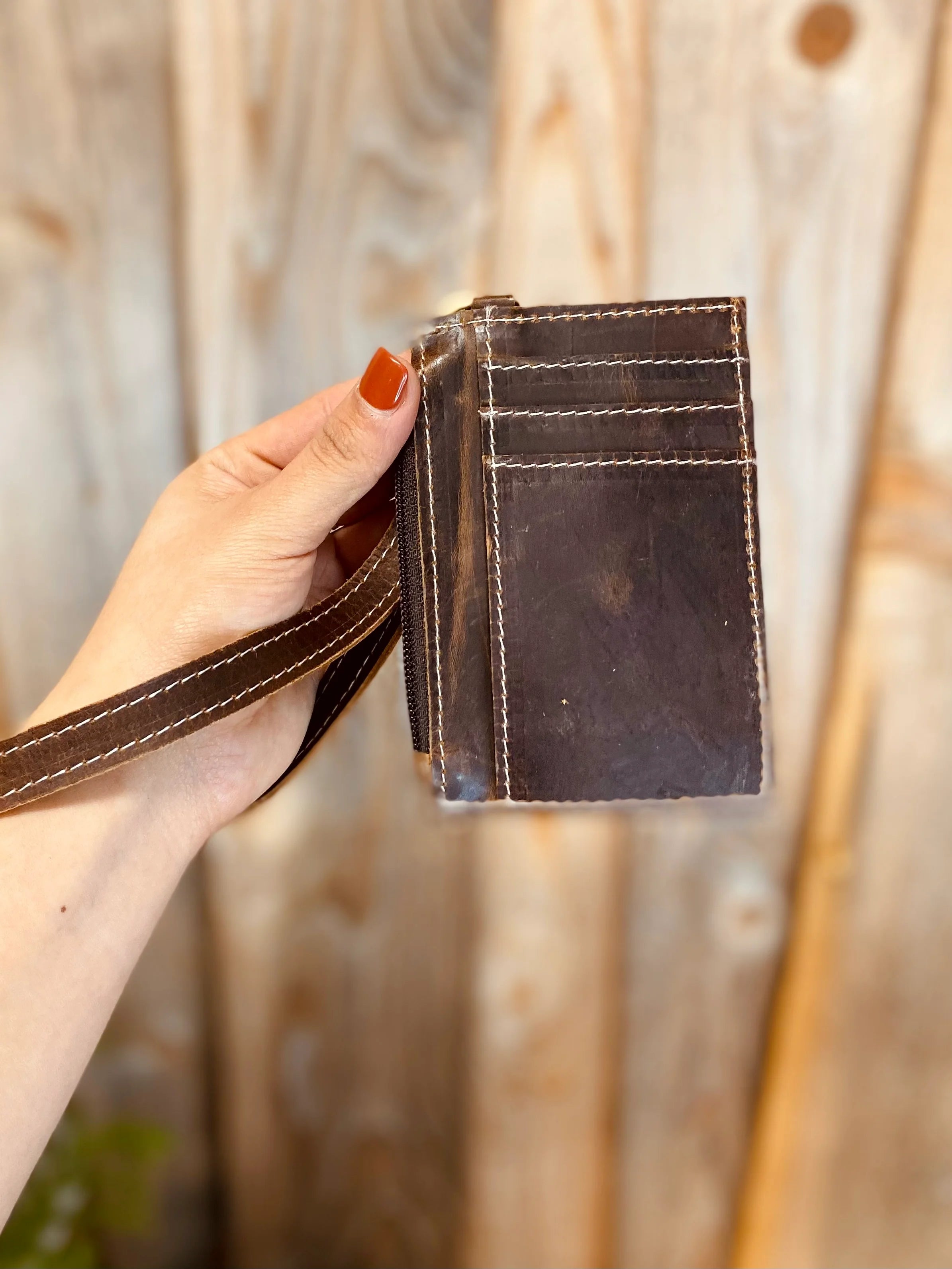 A person's hand holding a brown leather cardholder wristlet with a zipper pouch, against a wooden backdrop.
