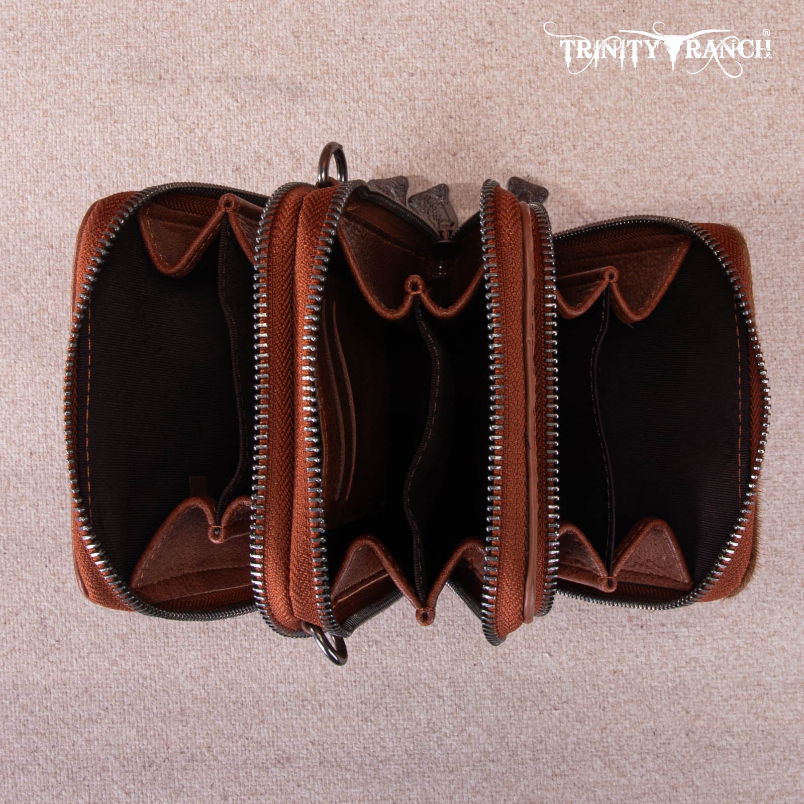 Brown leather wallet with zippers on a beige background, featuring 'Trinity Ranch' branding.