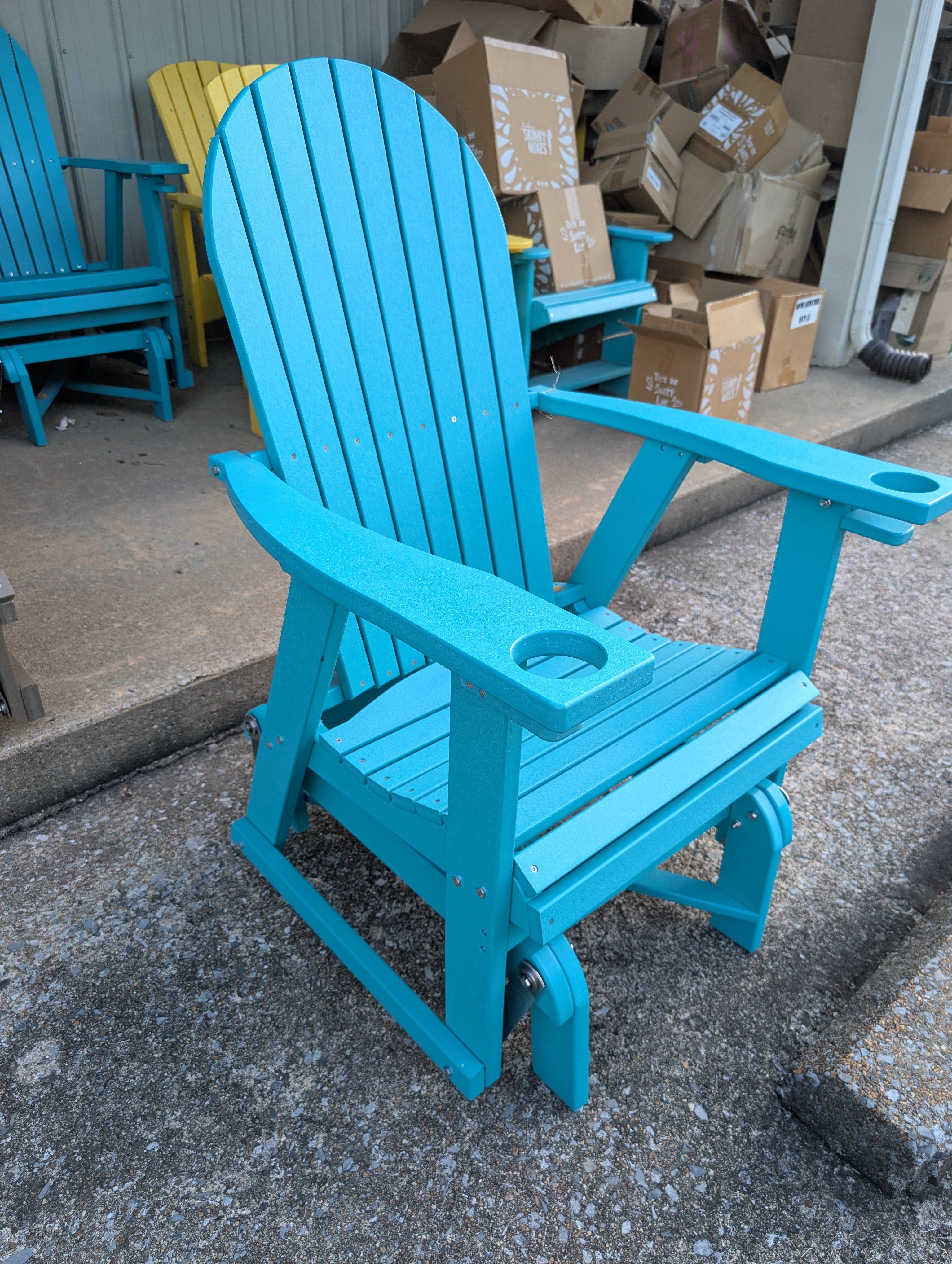 Blue Adirondack chair with cup holder on a concrete surface, with cardboard boxes in the background.