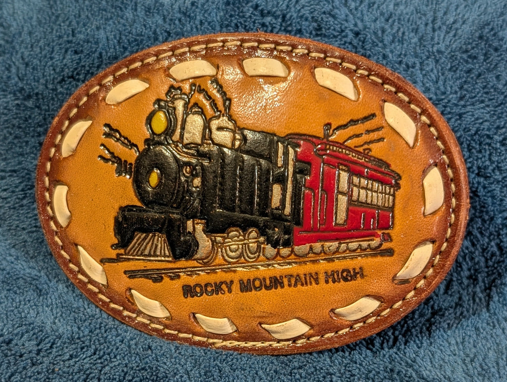 Leather belt buckle with train design and 'Rocky Mountain High' text on a blue textured background