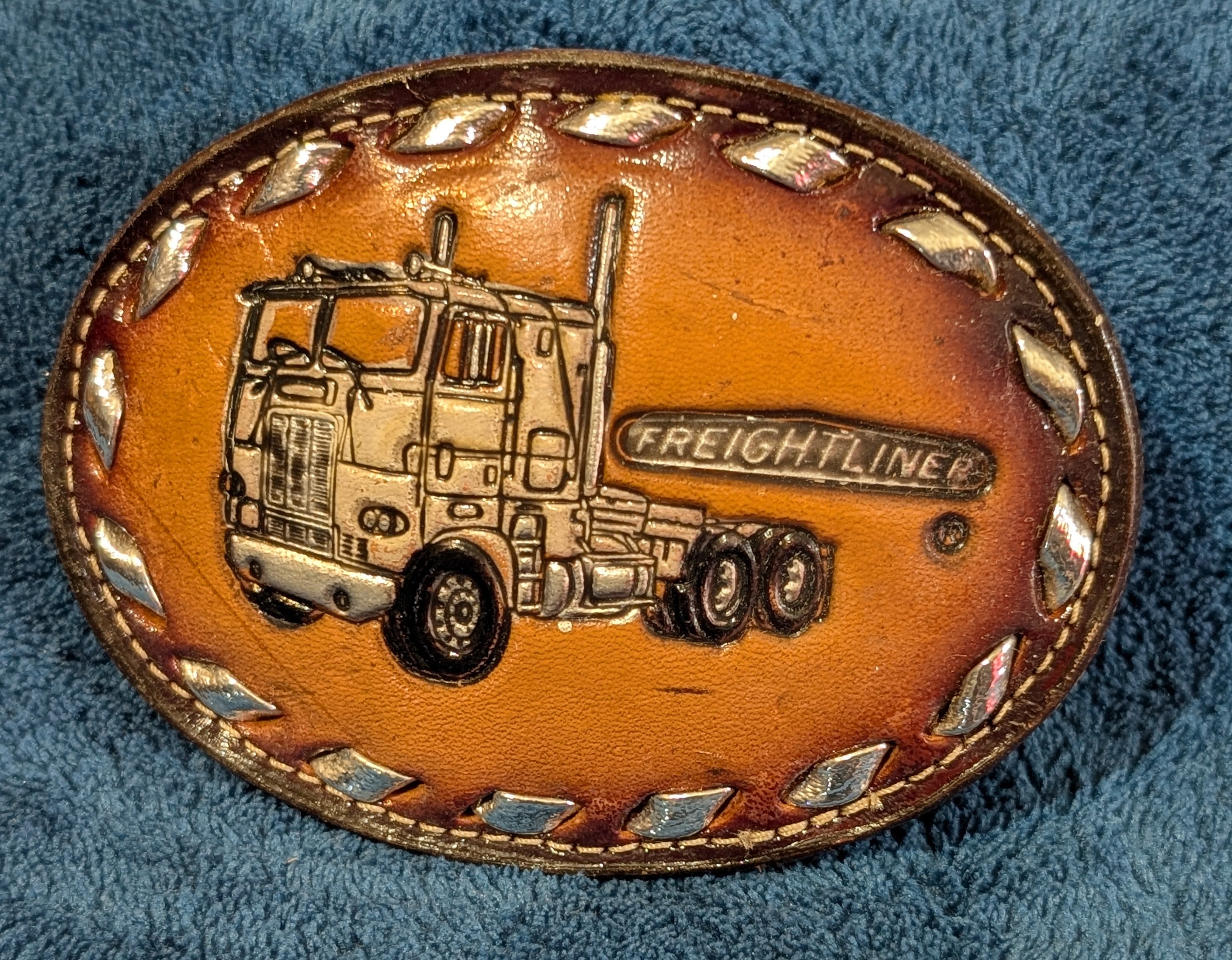 Leather belt buckle with truck design and 'Freightliner' branding on a blue fabric background