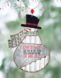 A festive snowman Christmas ornament hanging in front of a blurred evergreen tree. The ornament is made from galvanized tin with a distressed finish, featuring a black felt top hat with a red band, a green and red patterned scarf, and the text "CHRISTMAS THE TIME OF THE YEAR IT'S GOOD TO BE FAT AND JOLLY" in red and black.