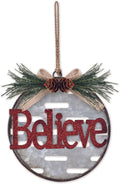 Oval metal Christmas ornament with the word "Believe" in glittery red script, hanging from a brown string. The ornament features a textured gray center, a rustic dark metal rim, a beige ribbon bow, pine green needles, and two small pinecones at the top.