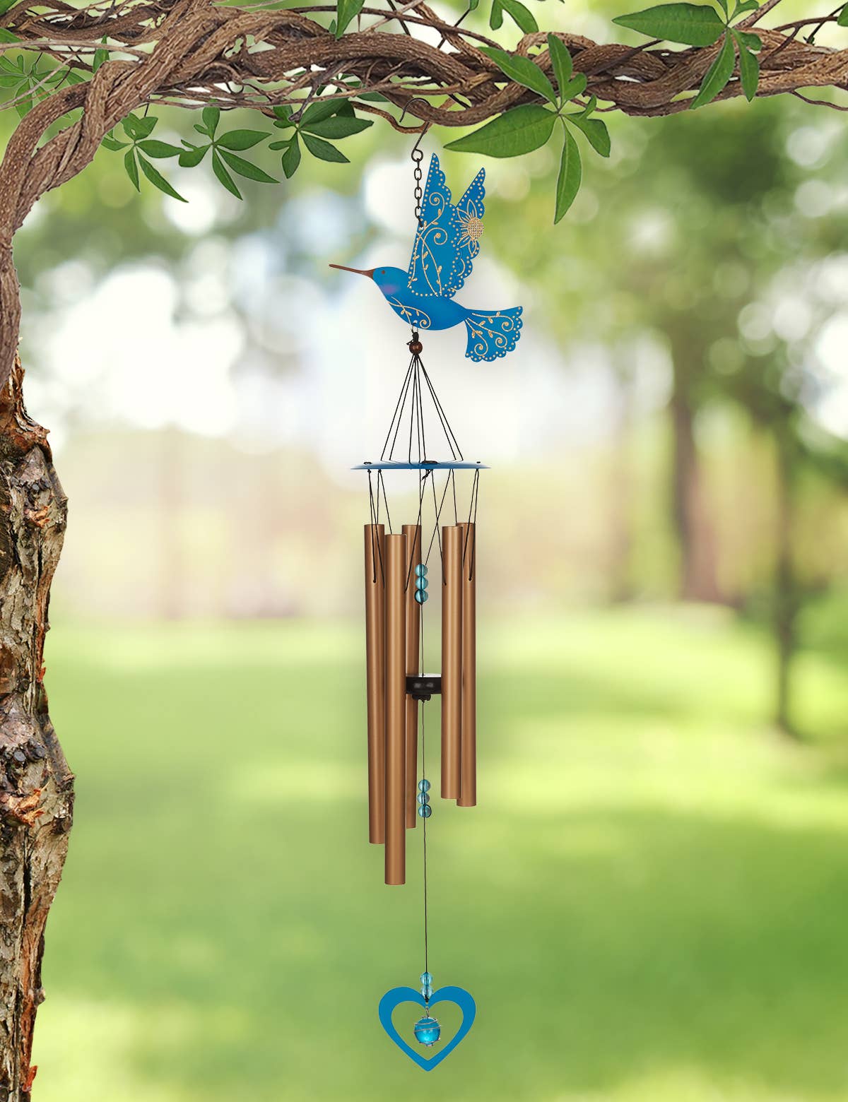 A blue hummingbird wind chime hanging from a tree branch in a garden. The chime features a blue hummingbird at the top, four brown metal tubes in the center, and a blue heart-shaped ornament at the bottom, set against a blurred green garden background.