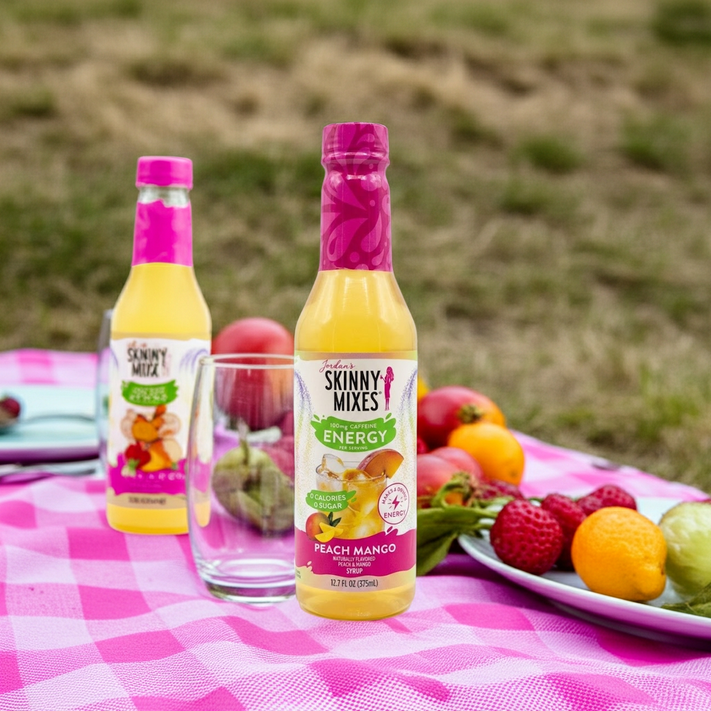 Two bottles of Jordan's Skinny Mixes Peach Mango flavor on a picnic blanket with fruits i