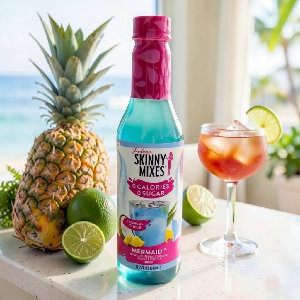 A bottle of Jordan's Skinny Mixes Mermaid tropical lime cocktail mix stands on a countertop beside a whole pineapple, two limes, and a pink drink in a coupe glass, with a bright beach background.