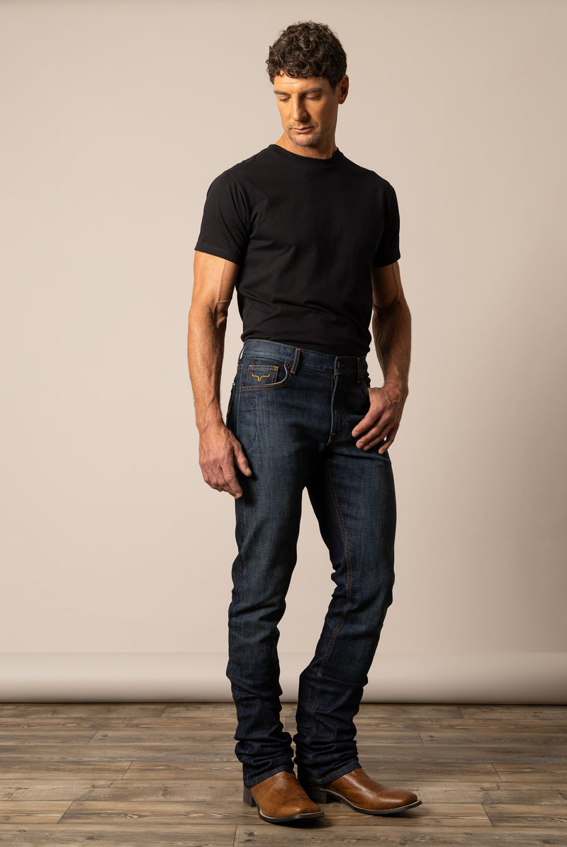 Man wearing a black t-shirt and Kimes Ranch dark jeans in the style Roger standing against a plain wall.