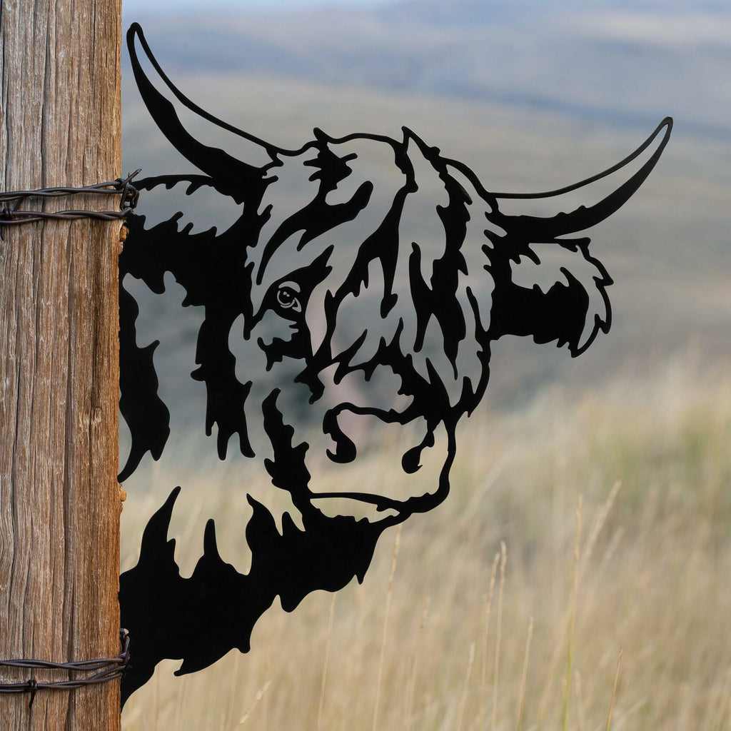 Rustic metal highland cow outdoor art from Homestead Collection peeking around wooden fence for farmhouse décor.