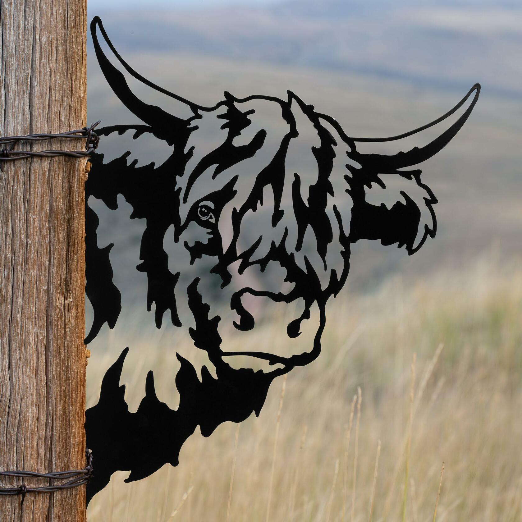 Rustic metal highland cow outdoor art from Homestead Collection peeking around wooden fence for farmhouse décor.