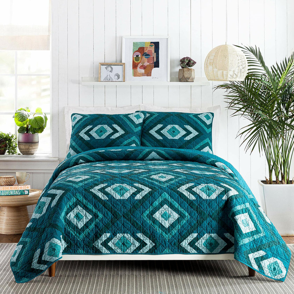 Teal diamond patterned quilt set on a bed in a bright bedroom with plants and decor.
