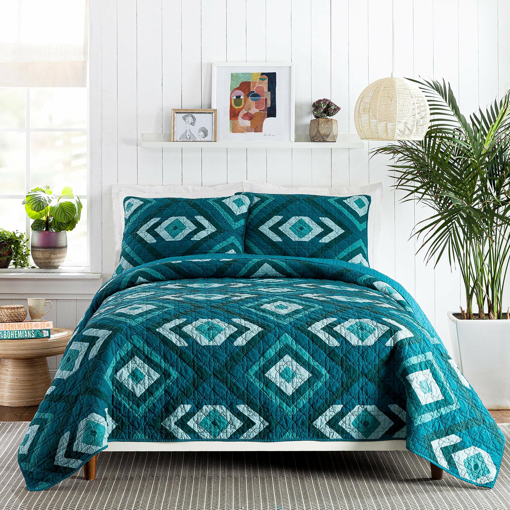 Teal diamond patterned quilt set on a bed in a bright bedroom with plants and decor.