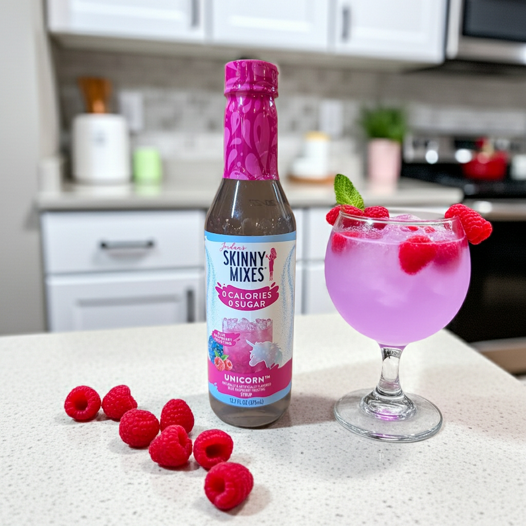 Jordan's Skinny Mixes Sugar-Free Unicorn Flavor Syrup 375ml bottle displayed on a white kitchen counter next to a purple mocktail garnished with fresh raspberries and mint.