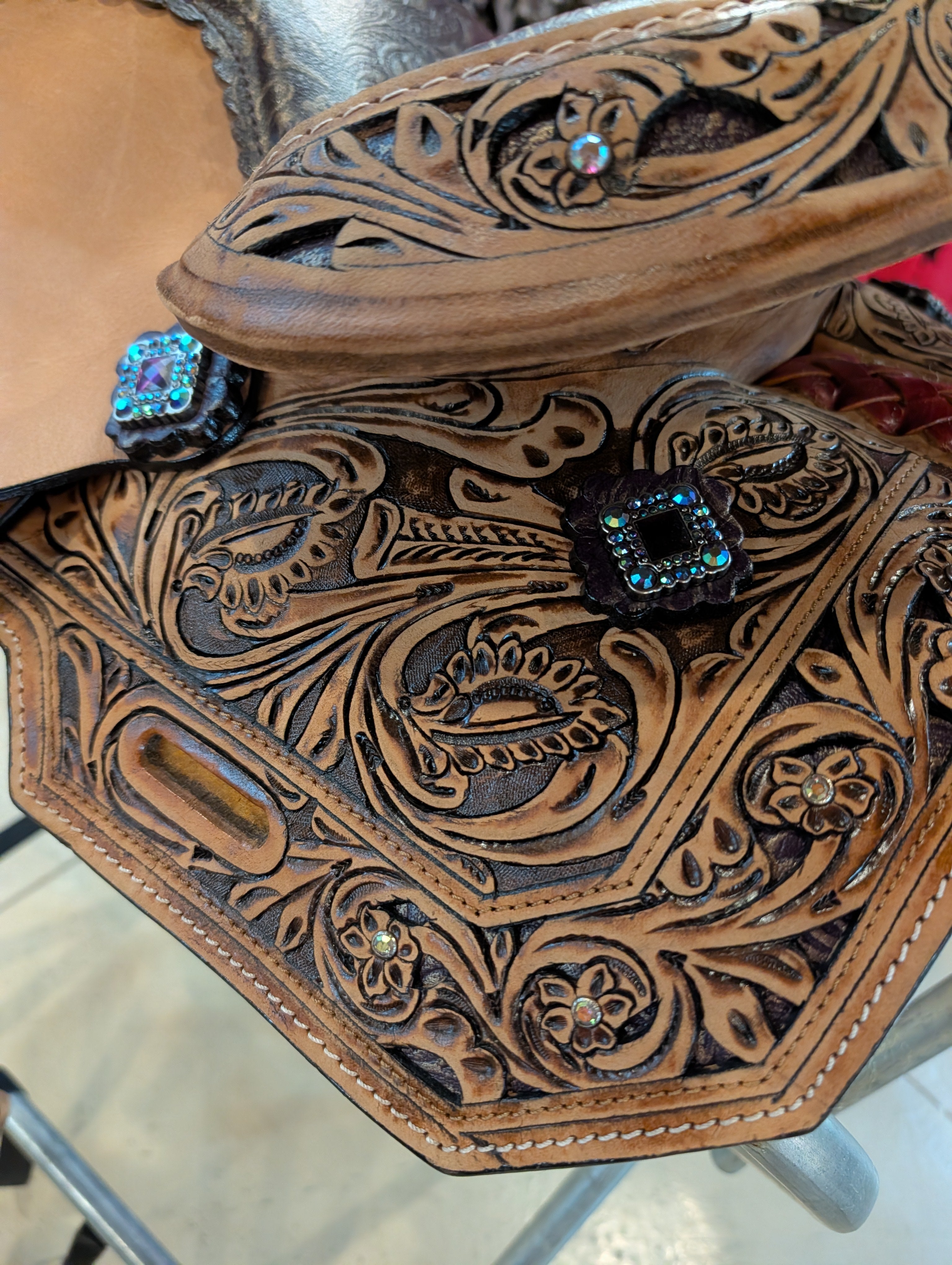 A close-up of a hand-tooled brown leather saddle or saddle-like western seat with ornate floral carving and turquoise rhinestone accents.